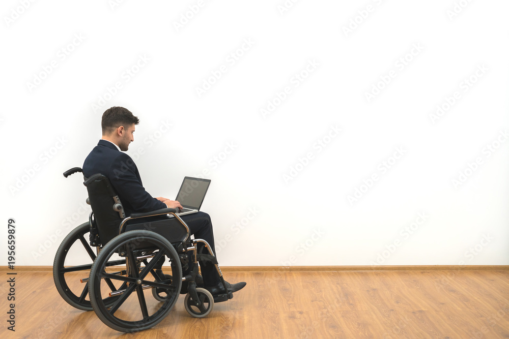 The man in a wheelchair working with a laptop on the white wall background The man in a wheelchair working with a laptop on the white wall background