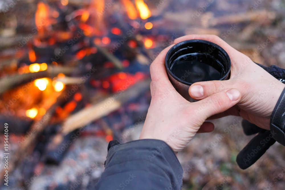Man hands holding mug from thermos near the fire outdoors.Traveler drinking tea from mug at camp Man hands holding mug from thermos near the fire outdoors.Traveler drinking tea from mug at camp