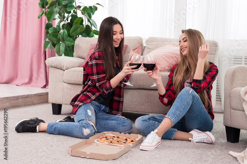 Time to relax! Two smart stylish girls   dressed in blue scruffy jeans with red plaid shirts clinking glasses with wine for a toast while sitting on the carpet at home.