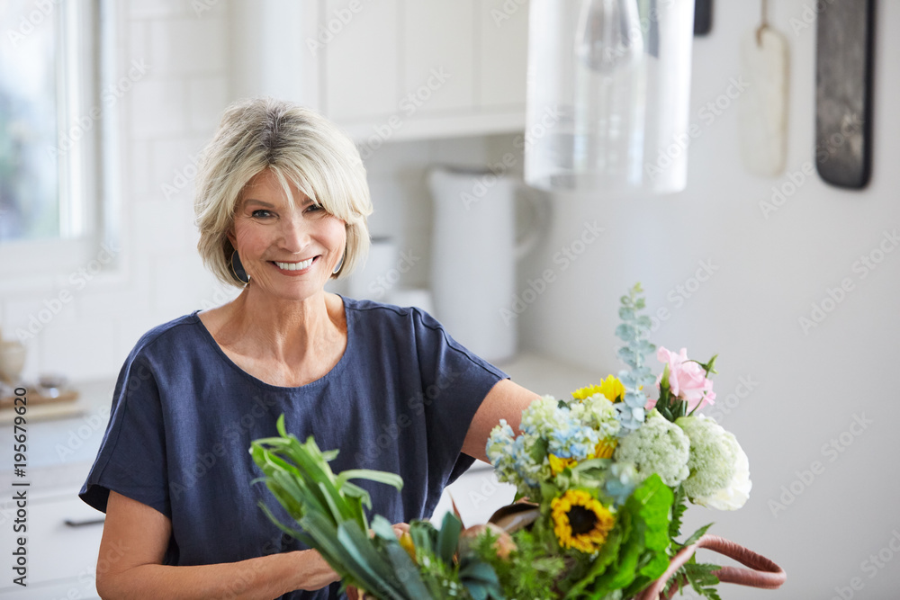 Senior woman in kitchen with farmers market basket with vegetables and flowers Senior woman in kitchen with farmers market basket with vegetables and flowers