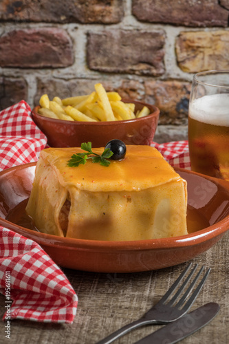 Traditional Portuguese snack food. Francesinha sandwich of bread cheese pork ham sausages with tomato beer sauce and french fries. With a glass of beer and potatoes. On wooden table.