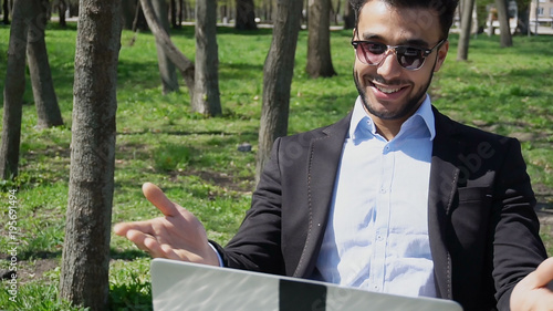 Handsome man sits by table, uses laptop and smiling with dimples on cheeks. Guy has white teeth, short dark hair and beard. Concept of apps for talking new lovers and modern technologies.