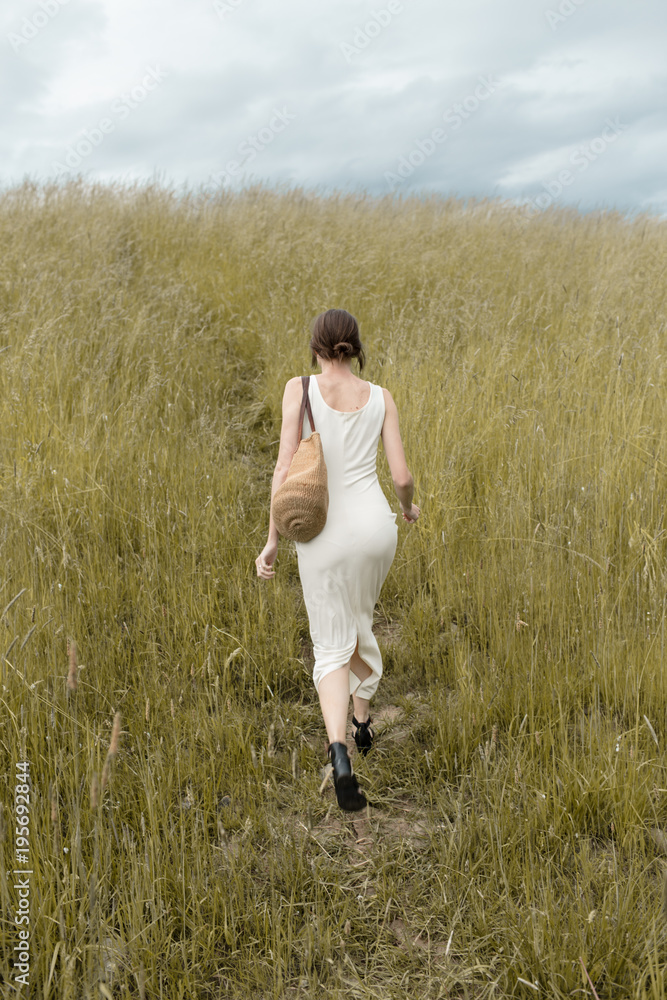 woman in a white dress carrying a bag walking up a grassy hill woman in a white dress carrying a bag walking up a grassy hill