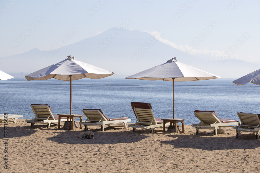 Tropical sand beach with sun loungers and umbrellas with a view of the volcano next to the sea water in island Bali, Indonesia