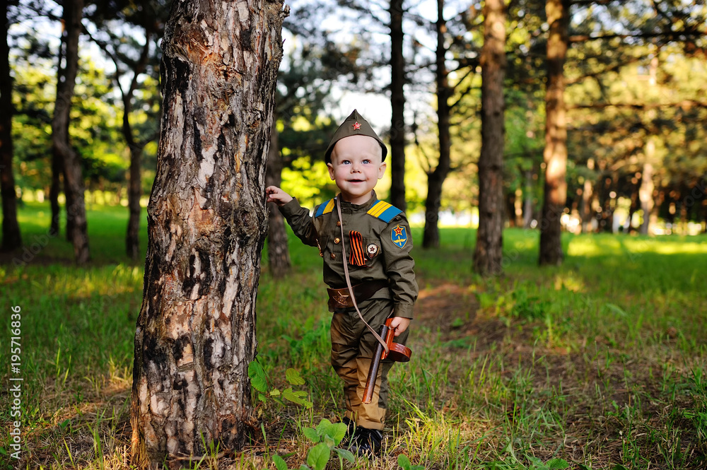 A cute child in the uniform of a Soviet soldier with a St. George ribbon with a toy gun on the background of nature. May 9, February 23, defender, against fascism. A cute child in the uniform of a Soviet soldier with a St. George ribbon with a toy gun on the background of nature. May 9, February 23, defender, against fascism.