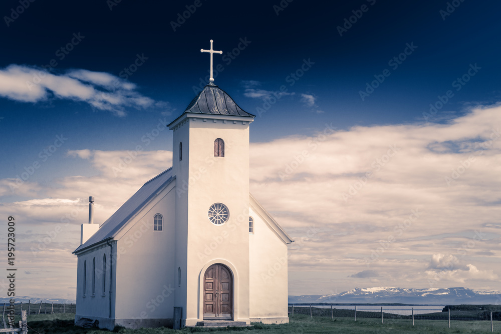 Flateyjarkirkja white lutheran church with meadow in foreground and sea  fjord with dark blue sky and mountains in the background, Flatey, Iceland