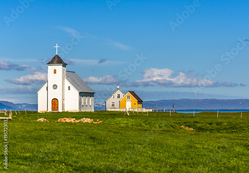 Flateyjarkirkja white lutheran church and couple of living huts with meadow in foreground and sea  fjord with blue sky in the background, Flatey, Iceland photo
