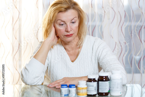 Portrait of 60-65 years old woman in white sweater sitting at the table and looking at medicine bottles. Medical health concept.