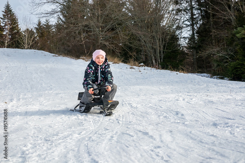 Winter snow activity. Front view of a girl riding a sledge downhill surrounded by trees.