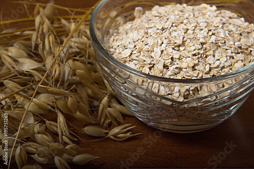 Oat ears stems and oat flakes in a bowl on a dark brown wood background. oat flakes small size grind. Close up. Useful fiber-rich product. Dietary breakfast from healthy foods