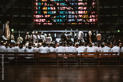 Rear view of the crowd of novices in white shirts sitting on the long wooden benches in a dark Catholic cathedral interior during a Christmas liturgy with a solemn ceremony in the defocused background photo