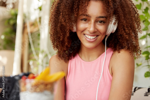 Afro American female student listens songs from playlist, enjoys perfect sound in modern headphones, has cheerful expression, sits against cafe interior. People, leisure, entertainmet concept photo