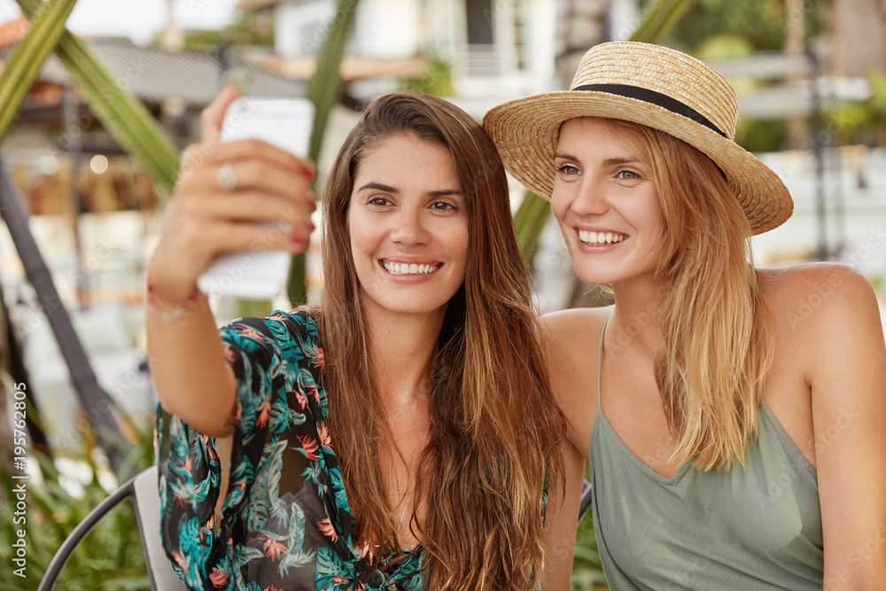 Happy beautiful females with broad smiles pose for selfie while sit together in cozy exotic cafteria, use modern smart phone for making photo. Cheerful lesbains make video call online via cellular