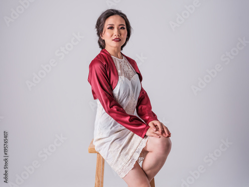 Beautiful Female Asian Model in white dress and red jacket sitting on wooden stool photo