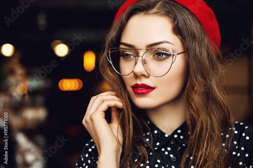 Close up portrait of young beautiful woman wearing stylish glasses, red beret, polka dot blouse. Model looking aside. Lights on background. Copy, empty space for text