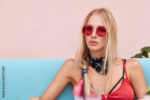 Portrait of pleasant looking pinup girl wears red stylish shades, has serious look, recreats on comfortable blue sofa, enjoys good rest. Blonde fashionable female feeles relaxed at cafeteria.