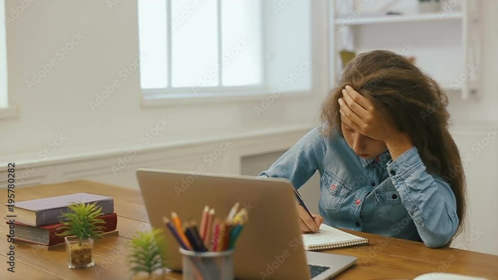 Young girl with laptop computer is doing homework. College education. Tired Female student studying and using pc at home. Young girl with laptop computer is doing homework. College education. Tired Female student studying and using pc at home.