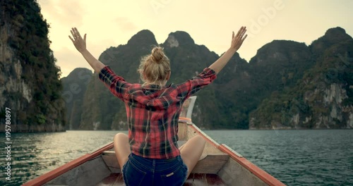 Back view of woman sitting in wood long tail boat with raised hands over tropical limestone cliffs and sunset background - video in slow motion