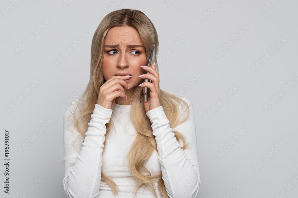 Woman nervous, bites finger, talking on mobile phone, frustrated due to an awkward situation, feeling shame, isolated on gray background. Worrying phone talk concept Woman nervous, bites finger, talking on mobile phone, frustrated due to an awkward situation, feeling shame, isolated on gray background. Worrying phone talk concept