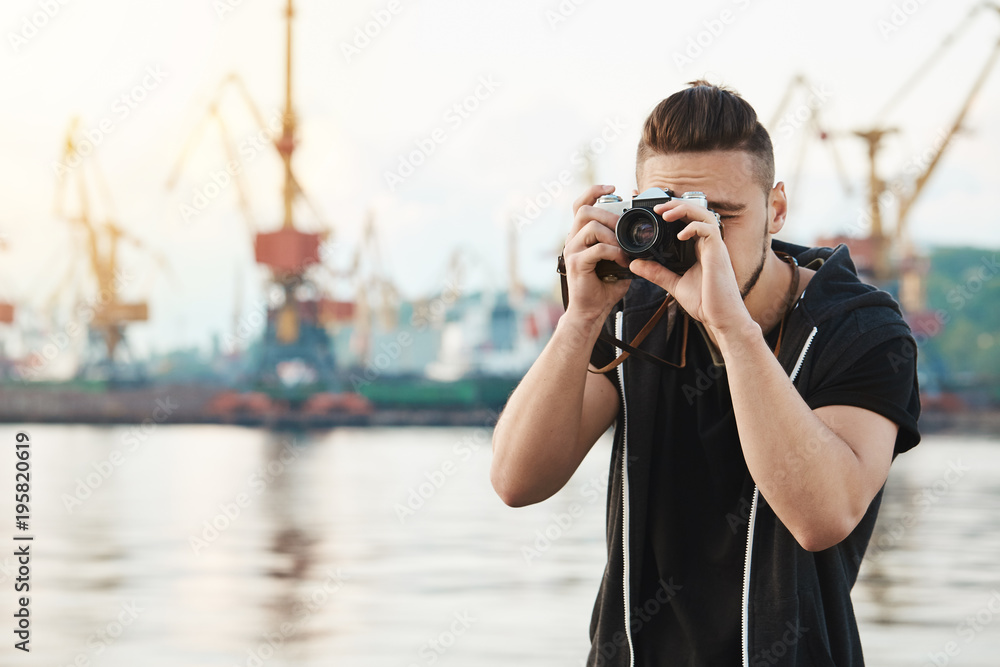 Attractive guy working with camera. Young stylish photographer looking through camera during photo session with gorgeous model, taking pictures in harbour near seashore, focusing on work