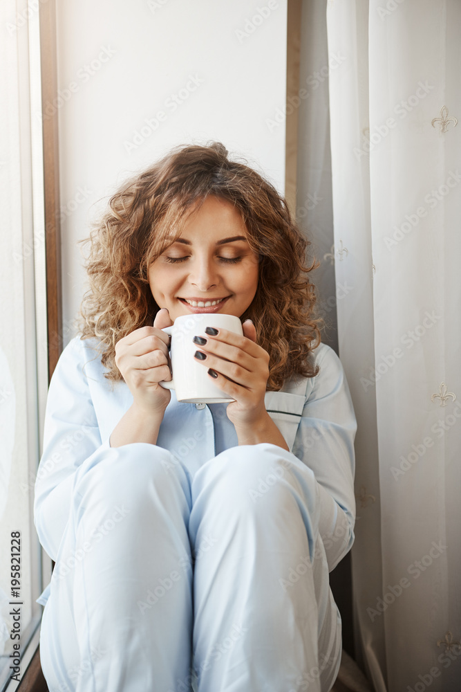 Vertical shot of beautiful caucasian young woman with curly hair sitting on window sill, holding cup of tasty coffee, smelling it with pleased and relaxed smile, having perfect morning alone Vertical shot of beautiful caucasian young woman with curly hair sitting on window sill, holding cup of tasty coffee, smelling it with pleased and relaxed smile, having perfect morning alone