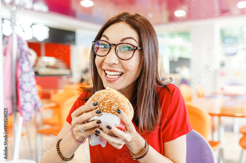 Happy healthy woman sitting in indoors food court and eating an delicious hamburger  modern meal concept