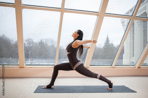 middle aged woman doing yoga in Virabhadrasana One or Warrior One yoga pose on the mat in front of large windows., exercise fitness, sport training, healthy lifestyle and people concept.