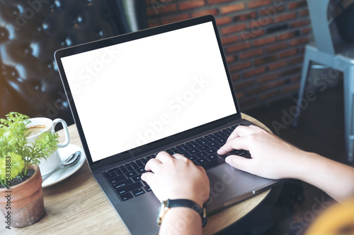 Mockup image of hands using and typing on laptop with blank white desktop screen and coffee cup on wooden table in cafe