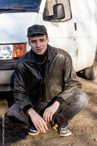 A young guy of criminal appearance in a black leather jacket stands near an old white van
