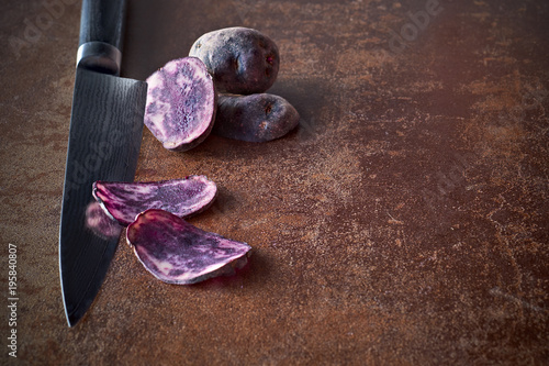 Sliced purple potatos, two slices on knife and two whole potatos on rough dark background photo