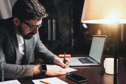 Bearded businessman sitting in night loft office and working with documents and contemporary laptop. Stylish man with glasses sits by the wooden table with lamp and write new plan