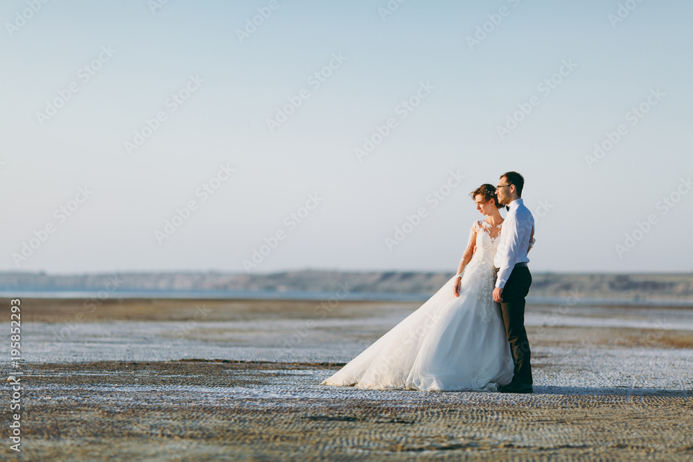 Beautiful wedding photosession. Handsome unshaved groom in a black trousers and young cute bride in white lace pattern dress with exquisite hairstyle on walk along the coastline near the sea sunset Beautiful wedding photosession. Handsome unshaved groom in a black trousers and young cute bride in white lace pattern dress with exquisite hairstyle on walk along the coastline near the sea sunset
