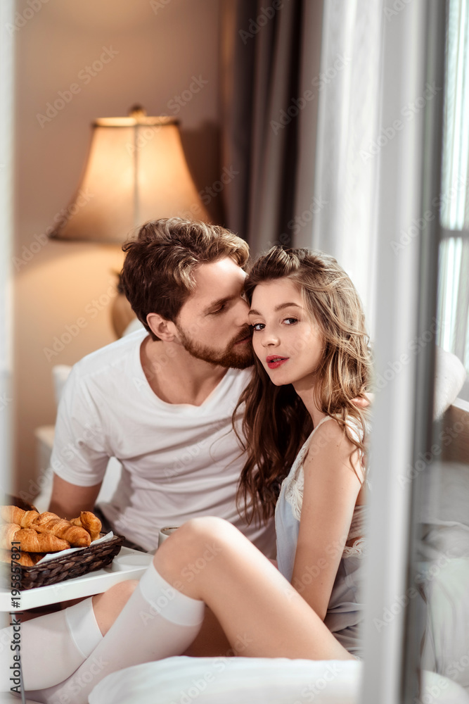 Vertical shot of pleasant looking female and her handsome bearded boyfriend who kisses her in cheek, enjoy together delicious croissants, sit in luxury hotel room, recreat abroad during vacation