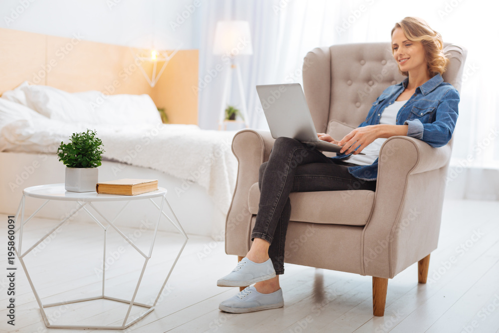 Productive day. Cheerful calm young woman sitting comfortably in a big soft armchair and feeling good while working on her modern laptop Productive day. Cheerful calm young woman sitting comfortably in a big soft armchair and feeling good while working on her modern laptop