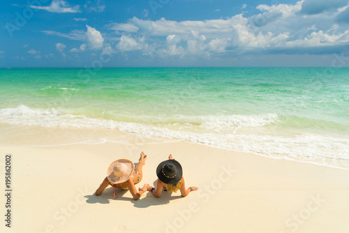 Fototapeta Naklejka Na Ścianę i Meble -  Sexy bikini body of two women enjoy the sea by laying down on sand of beach wearing hat. Happy island lifestyle. White sand and crystal sea of tropical beach.