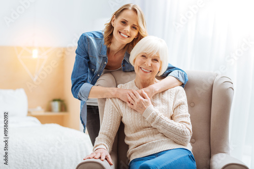 Close people. Positive emotional elderly woman sitting in a soft comfortable armchair while her kind attentive loving granddaughter standing behind her back and softly hugging her