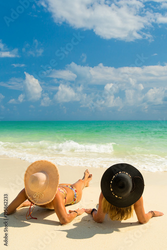 Sexy bikini body of two women enjoy the sea by laying down on sand of beach wearing hat. Happy island lifestyle. White sand and crystal sea of tropical beach.