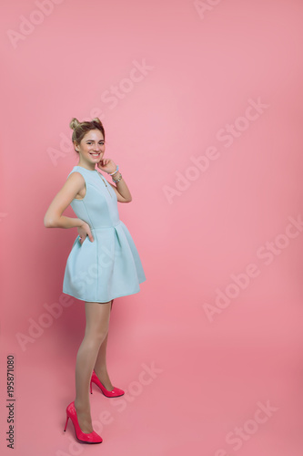 pretty youth woman wearing turquoise dress and high heels shoes, looking at camera and smiling, while standing against pink pastel background. Summer look for young woman