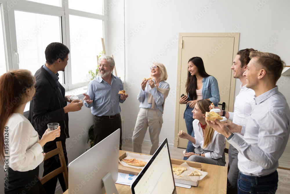 Friendly diverse business team of young and senior colleagues eating pizza together in office, multiracial coworkers staff group talking and laughing at funny joke on lunch time or coffee break
