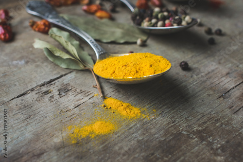 Close up view of a vintage spoon filled with curcuma or turmeric, with allspice pepper and spilled spices in the background on old rustic wooden background. Vintage toned. Spices background