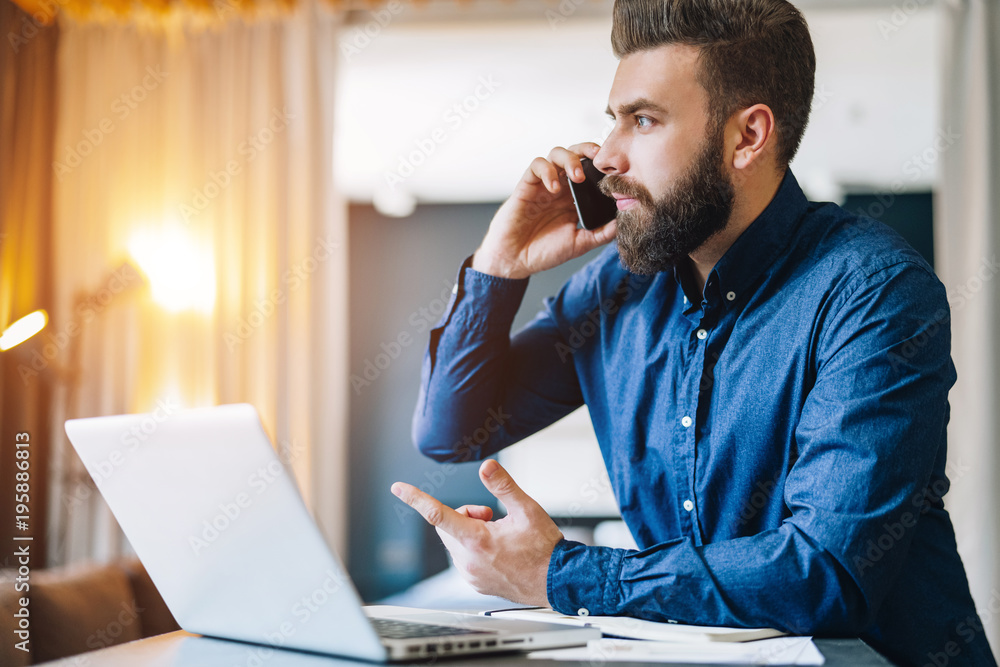 Young bearded businessman is sitting at table in front of computer, talking on cell phone. Freelancer, entrepreneur works at home. Telephone conversations. Distance work, online education, marketing. Young bearded businessman is sitting at table in front of computer, talking on cell phone. Freelancer, entrepreneur works at home. Telephone conversations. Distance work, online education, marketing.