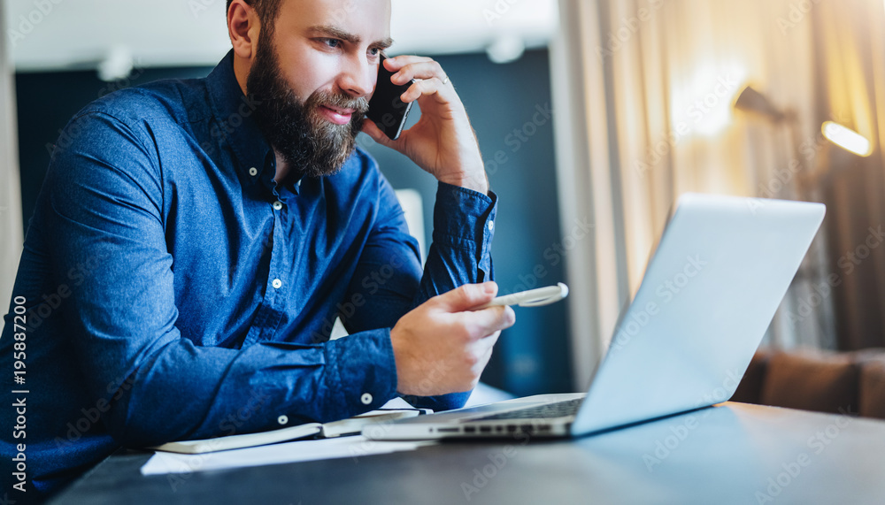 Young smiling bearded businessman is sitting at table in front of computer, talking on cell phone, showing pen on laptop screen. Freelancer works at home. Distance work, online education, marketing. Young smiling bearded businessman is sitting at table in front of computer, talking on cell phone, showing pen on laptop screen. Freelancer works at home. Distance work, online education, marketing.