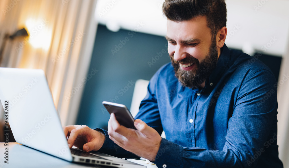 Young smiling bearded businessman sitting at table in front of computer, using smartphone. Freelancer, entrepreneur works at home. Online marketing, education for adults, distance work. Social network
