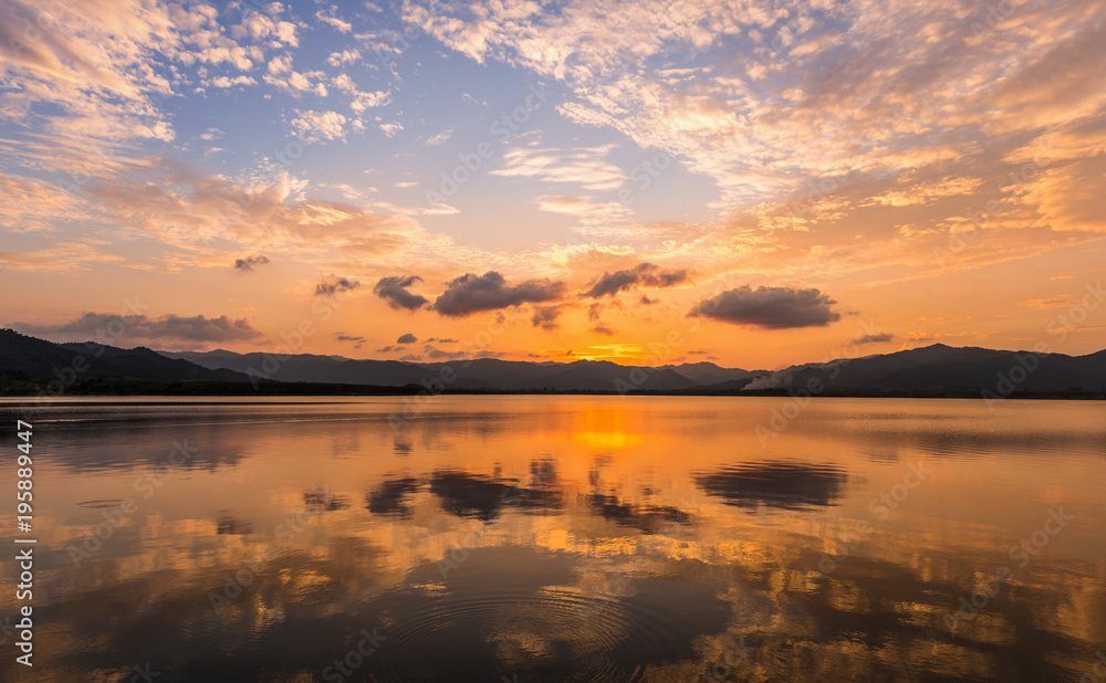 Twilight scene of clouds and golden sky reflecting on flat water surface of lake surounding by dark mountain ranges.