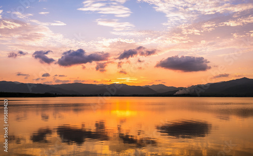 Twilight scene of clouds and golden sky reflecting on flat water surface of lake surounding by dark mountain ranges.