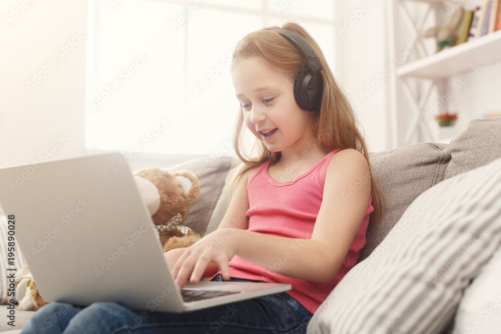 Cute little girl doing homework on laptop, sitting on couch in headphones Cute little girl doing homework on laptop, sitting on couch in headphones
