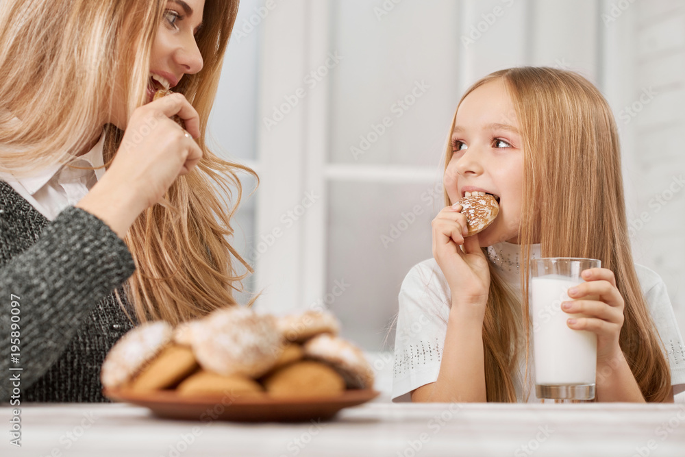 Photo of mother and daughter eating cookies and drinking milk. Both girls have blonde straight hair. They are eating delicious sweet cookies from plate and drinking milk near white kitchen's table.