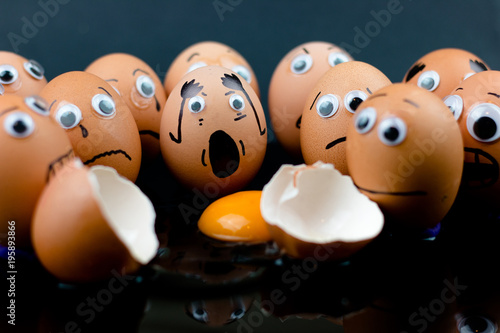 Vászonkép Fun concept: raw eggs with googly eyes and drawn features are in shock and sad as they witness another egg broken in two with the white and yolk in front of them; dark background