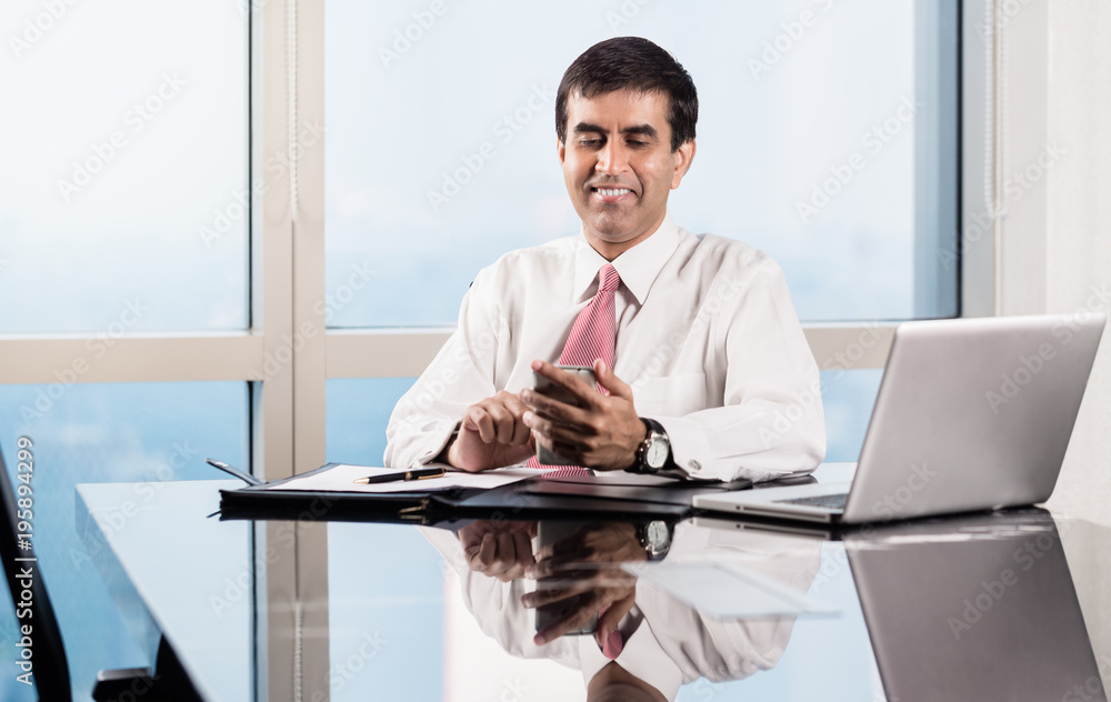Indian Senior business executive sitting at desk in skyscraper office checking his smart phone Indian Senior business executive sitting at desk in skyscraper office checking his smart phone