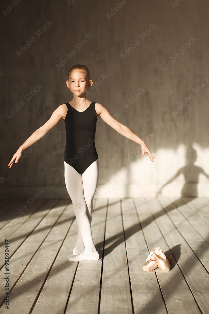 Naklejka premium A little adorable young ballerina in black sportwear and white tights exercising in the interior studio posing on camera in bright sunlight from window